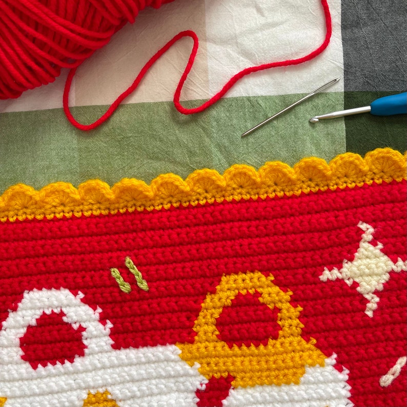 May include: Close-up of a red, white, and yellow crocheted blanket with a scalloped edge. The blanket features a design of flowers and leaves.