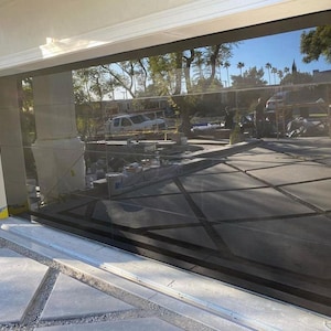 May include: A modern garage door with a dark glass panel and a sleek, minimalist design. The door is closed and reflects the surrounding landscape, including trees and a building in the distance.