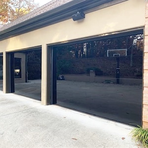 May include: Two black garage doors with glass panels, reflecting the surrounding trees and a basketball hoop. The doors are set against a beige wall with a brown trim.