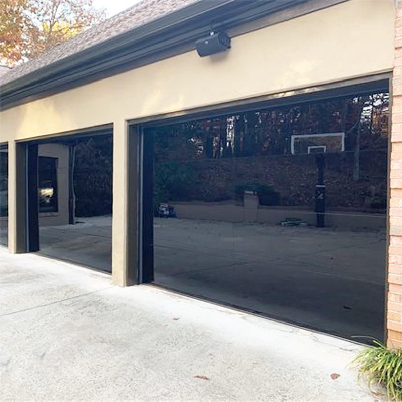 May include: Two black garage doors with glass panels, reflecting the surrounding trees and a basketball hoop. The doors are set against a beige wall with a brown trim.