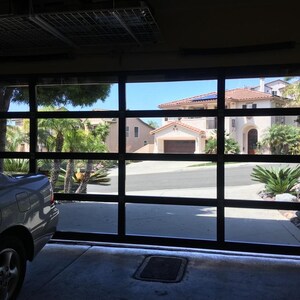 May include: A white car parked in a garage with a glass garage door. The garage door is open and you can see a house with a red tile roof and a driveway in the background.