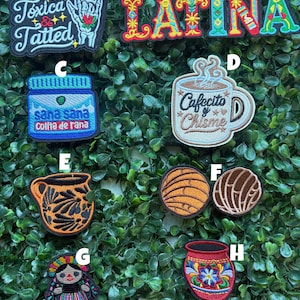 May include: Collection of embroidered patches with various designs. Includes patches with the text "Tóxica & Tatted", "Latina", "Cafecito y Chisme", and "sana sana colita de rana". Other patches feature a coffee mug, a traditional doll, and a decorative vase.