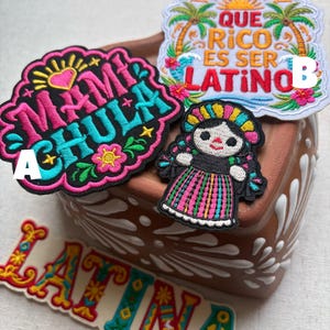 May include: Four embroidered patches featuring colorful designs. Patch A reads MAMI CHULA in bright pink and teal. Patch B reads QUE RICO ES SER LATINO with palm trees and sun. Patch C reads LATINA in decorative letters. A small doll patch is also included.