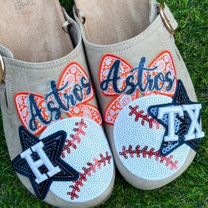 May include: A pair of beige slip-on sandals featuring orange paisley bow patches, white sequined baseball patches with red stitching, and dark navy star patches. One sandal displays an H and the other a TX, with the word Astros in glitter.