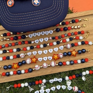 May include: A navy blue baseball cap with an orange and white logo, surrounded by beaded bracelets. The bracelets feature red, white, and blue beads, baseball charms, and letter beads spelling "ASTROS", "HTX", and "HOUSTON".