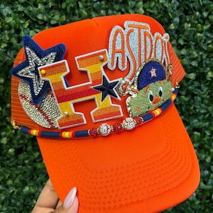 May include: Orange trucker hat with the word "ASTROS" in silver glitter and a cartoon character. The hat features a star and baseball design, with a beaded band around the brim. The hat is a vibrant orange color.