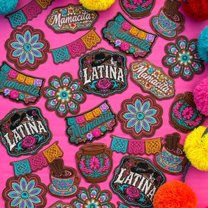 May include: A collection of embroidered patches featuring various designs including flowers, cups of hot chocolate, papel picado banners, and text reading Mamacita or Latina, accented by colorful yarn pom-poms on a bright pink background.
