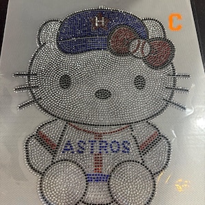 May include: A rhinestone transfer design featuring a cartoon cat wearing a blue baseball cap with an H logo and a baseball-patterned bow. The cat wears a white jersey with the text ASTROS in blue letters. The design is made of clear, blue, and red rhinestones.