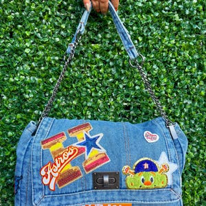 May include: A light blue denim handbag featuring a metallic chain strap and a turn-lock closure. The bag is decorated with various embroidered patches, including a large Astros logo, a cartoon character, and a patch with the text BE SOMEONE.