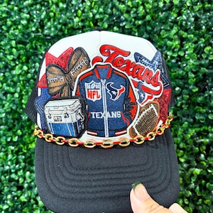 May include: A white and black trucker hat featuring embroidered patches of a Houston Texans varsity jacket, a Yeti cooler, footballs, and red and blue stars. A gold chain with a red accent line is attached across the base of the front panel.