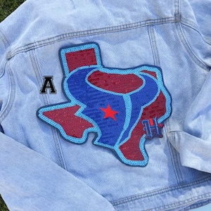 May include: Light blue denim jacket with a large Texas-shaped patch on the back. The patch features a red and blue design with a star and the letters "A" and "H". The jacket has a classic design with a button closure and pockets.