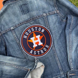 May include: A blue denim jacket with a large Houston Astros patch on the back. The patch features a navy blue background with the team name and logo in orange and white. The jacket is made of a medium-wash denim.
