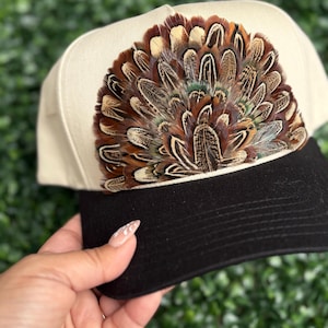 May include: A cream-colored baseball cap with a black brim, adorned with a fan of brown, tan, and green feathers. The feathers are arranged in a semi-circle, creating a unique and eye-catching design. This hat is a stylish accessory.