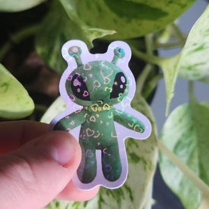 Op de afbeelding: Een holografische sticker van een groene alien met grote ogen en een hartpatroon. De alien draagt een gouden ketting en heeft een witte achtergrond.