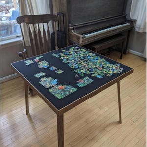 May include: A square wooden puzzle table with a black felt surface. The table has four angled legs and is partially covered with a colourful jigsaw puzzle. The table is in a room with a piano and a wooden chair.