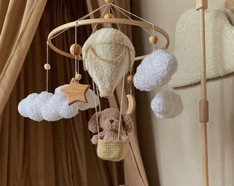 May include: A wooden baby mobile with a brown teddy bear in a basket, a white hot air balloon, white clouds, a wooden star, and a crescent moon. The mobile is hanging from a wooden stand.