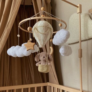 May include: A wooden baby mobile with a brown teddy bear in a basket, a white hot air balloon, white clouds, a wooden star, and a crescent moon. The mobile is hanging from a wooden stand.