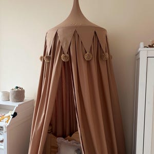 May include: A tan-colored bed canopy with a conical top and draped sides. The canopy is decorated with triangular accents and fluffy pom-poms. The canopy is suspended above a bed, creating a cozy and decorative space.