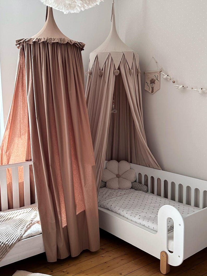 Pu&ograve; includere: Due letti per bambini con baldacchini in una palette di colori neutri. Un baldacchino ha un bordo a balze, l'altro ha accenti triangolari e pompon. I letti hanno telai bianchi e uno ha un cuscino a forma di fiore.