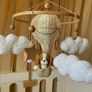 May include: A wooden baby mobile with a hot air balloon, clouds, a star, and a dog in a basket. The balloon is beige and white, the clouds are white, the star is wooden, and the dog is brown and white.