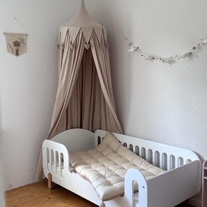Pu&ograve; includere: Un lettino bianco con un materasso e un cuscino trapuntati beige. Una tenda da letto beige &egrave; appesa sopra il letto, adornata con dettagli a forma di cuore. La stanza presenta un pavimento in legno e una parete con un motivo a pois.