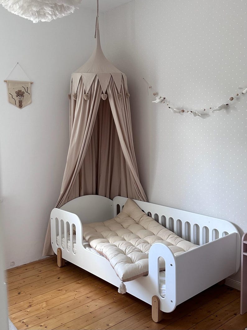 Pu&ograve; includere: Un lettino bianco con un materasso e un cuscino trapuntati beige. Una tenda da letto beige &egrave; appesa sopra il letto, adornata con dettagli a forma di cuore. La stanza presenta un pavimento in legno e una parete con un motivo a pois.