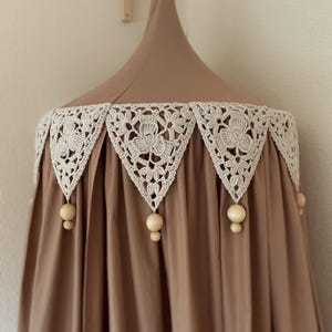 Pu&ograve; includere: Un baldacchino marrone con un bordo in pizzo all'uncinetto color crema con motivi floreali. Perline di legno pendono dal bordo. Il baldacchino &egrave; progettato per essere appeso al soffitto, creando un effetto morbido e drappeggiato.