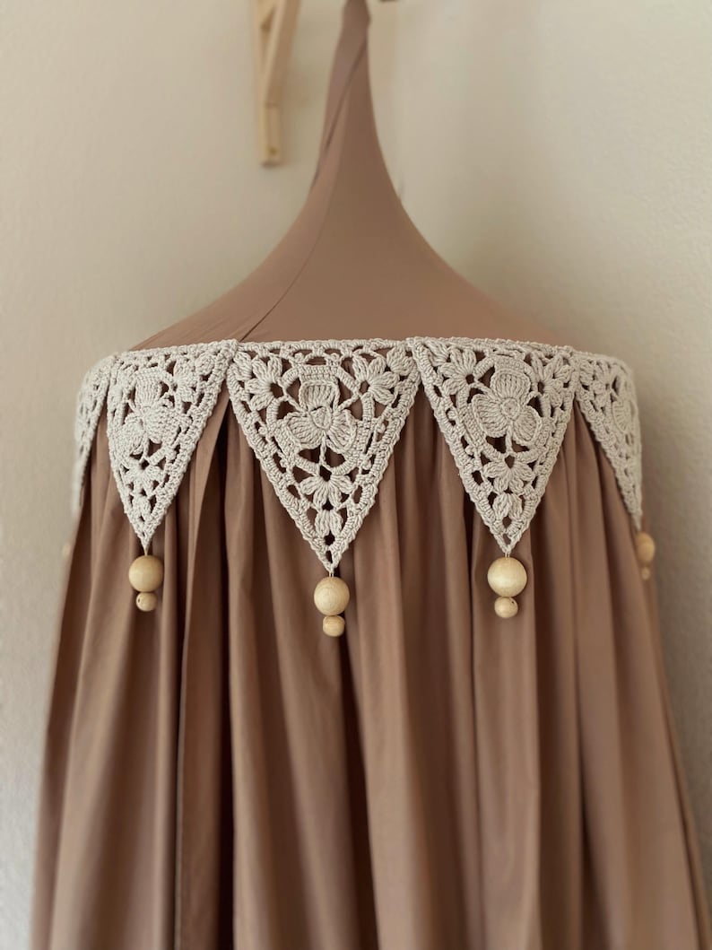 Pu&ograve; includere: Un baldacchino marrone con un bordo in pizzo all'uncinetto color crema con motivi floreali. Perline di legno pendono dal bordo. Il baldacchino &egrave; progettato per essere appeso al soffitto, creando un effetto morbido e drappeggiato.