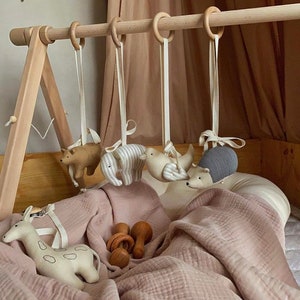 May include: A wooden baby play gym with hanging toys, including a giraffe, elephant, bird, hedgehog, and bear. The gym is set up on a pink blanket with two wooden teethers.