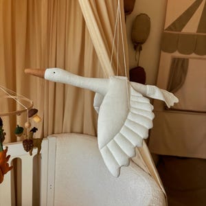 May include: A white fabric swan mobile hanging from the ceiling above a white crib. The swan has a brown beak and is made of soft, quilted fabric.