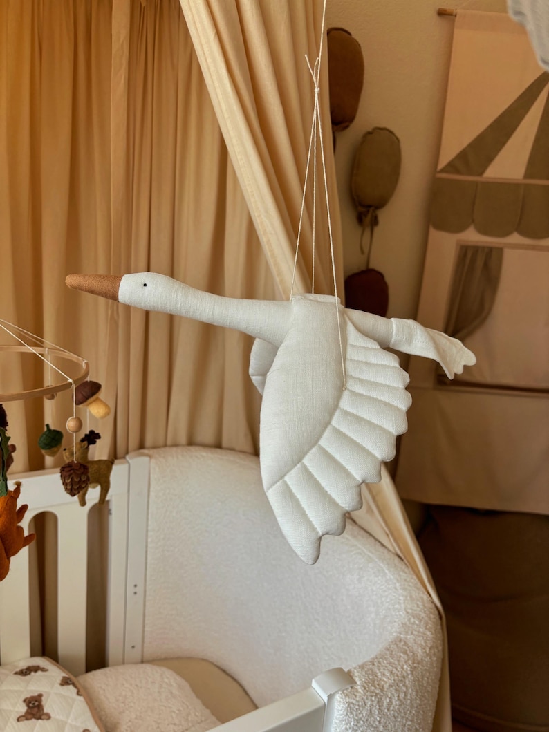 May include: A white fabric swan mobile hanging from the ceiling above a white crib. The swan has a brown beak and is made of soft, quilted fabric.
