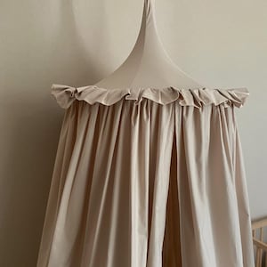 Pu&ograve; includere: Un baldacchino beige con un bordo a balze e una parte superiore conica. Il tessuto drappeggia verso il basso, creando un effetto morbido e fluido. Il baldacchino &egrave; sospeso da un singolo punto, suggerendo un elemento decorativo per la cameretta o la camera da letto.