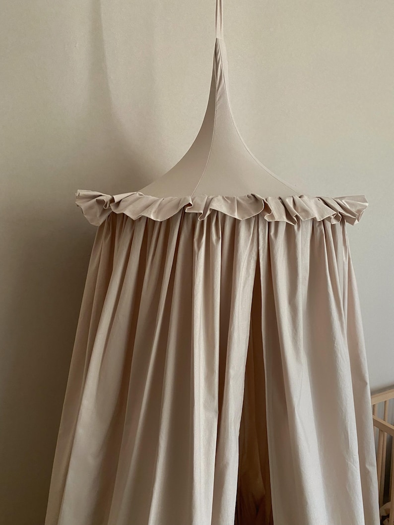 Pu&ograve; includere: Un baldacchino beige con un bordo a balze e una parte superiore conica. Il tessuto drappeggia verso il basso, creando un effetto morbido e fluido. Il baldacchino &egrave; sospeso da un singolo punto, suggerendo un elemento decorativo per la cameretta o la camera da letto.