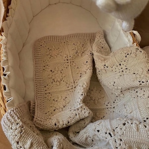 Op de afbeelding: Een collectie handgemaakte gehaakte babydekens in zachte beige en crèmetinten. De dekens hebben bloempatronen en decoratieve randen, gepresenteerd in een rieten wieg met een wit interieur. De algehele esthetiek is delicaat en gezellig.