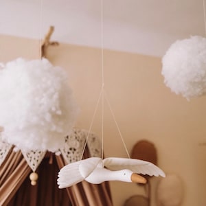 May include: A white wooden bird mobile hanging from the ceiling with white fluffy pom poms and a brown canopy in the background.