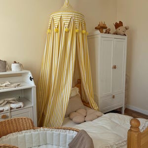 May include: A child's bedroom with a yellow and white striped canopy. A wicker bassinet, white bedding, and a white wardrobe are present. The room has a soft, neutral colour palette, creating a calming atmosphere.