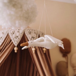 May include: A white fabric goose mobile hanging from the ceiling above a brown canopy bed. The goose has a brown beak and is suspended by white strings.