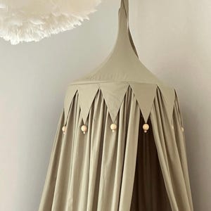 Pu&ograve; includere: Un baldacchino verde salvia con la parte superiore conica e accenti triangolari. Perline di legno pendono dal bordo inferiore. Sullo sfondo &egrave; visibile una nuvola decorativa bianca e soffice. La tenda &egrave; sospesa al soffitto.