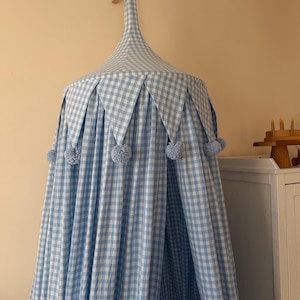 May include: A blue and white gingham canopy with a pointed top and pleated edges. The canopy is adorned with light blue pom-poms and is suspended from a wooden bracket. The fabric is a light blue and white checkered pattern.