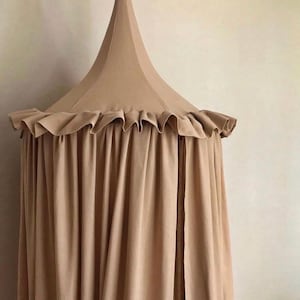 Pu&ograve; includere: Un baldacchino in tessuto beige con una parte superiore conica e finiture a balze. Il baldacchino pende in morbide pieghe, creando un effetto drappeggiato. Il colore neutro e il design semplice suggeriscono un arredamento per camerette o asili nido.
