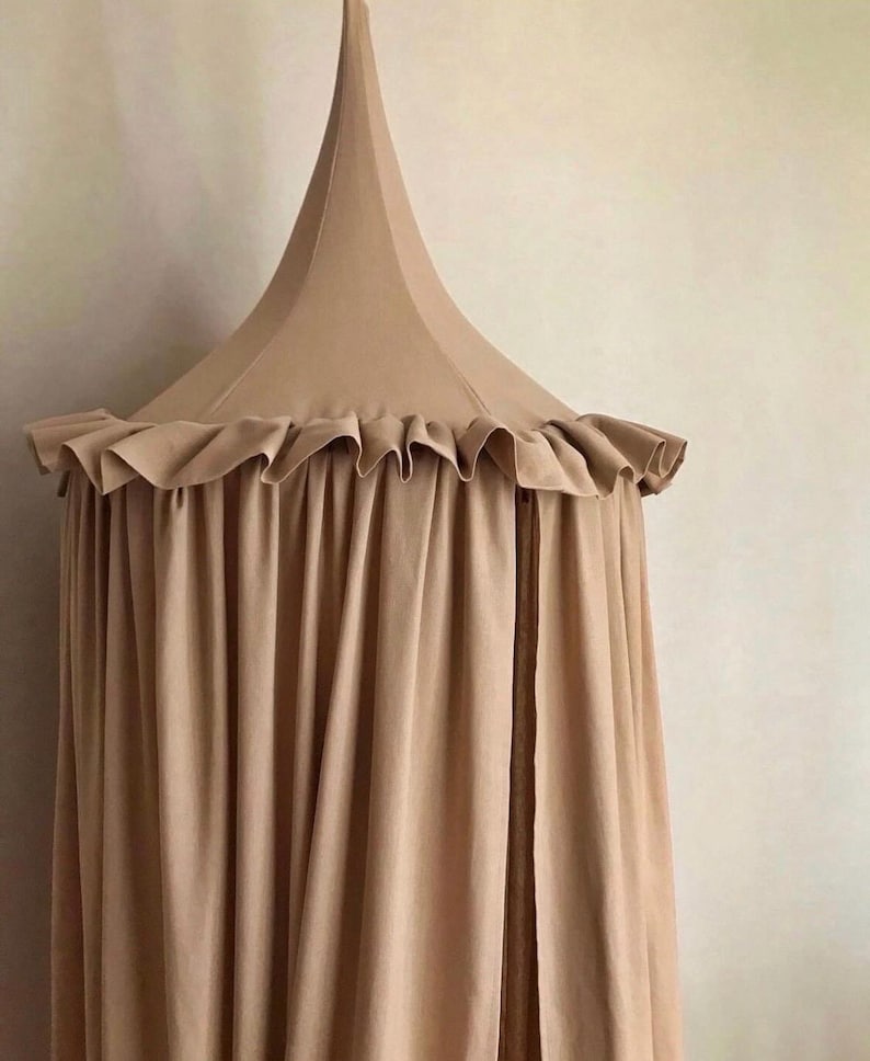 Pu&ograve; includere: Un baldacchino in tessuto beige con una parte superiore conica e finiture a balze. Il baldacchino pende in morbide pieghe, creando un effetto drappeggiato. Il colore neutro e il design semplice suggeriscono un arredamento per camerette o asili nido.