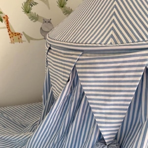 May include: A conical fabric canopy featuring a blue and white striped pattern. The canopy includes a decorative bunting border with triangular pennants, each finished with a small, round, light blue pom-pom at the bottom point of the fabric.