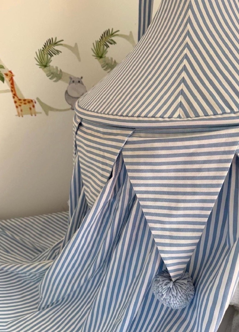 May include: A conical fabric canopy featuring a blue and white striped pattern. The canopy includes a decorative bunting border with triangular pennants, each finished with a small, round, light blue pom-pom at the bottom point of the fabric.