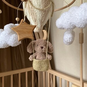 May include: A wooden baby mobile with a crocheted hot air balloon, clouds, and a star. A brown plush puppy sits in a crocheted basket hanging from the balloon.