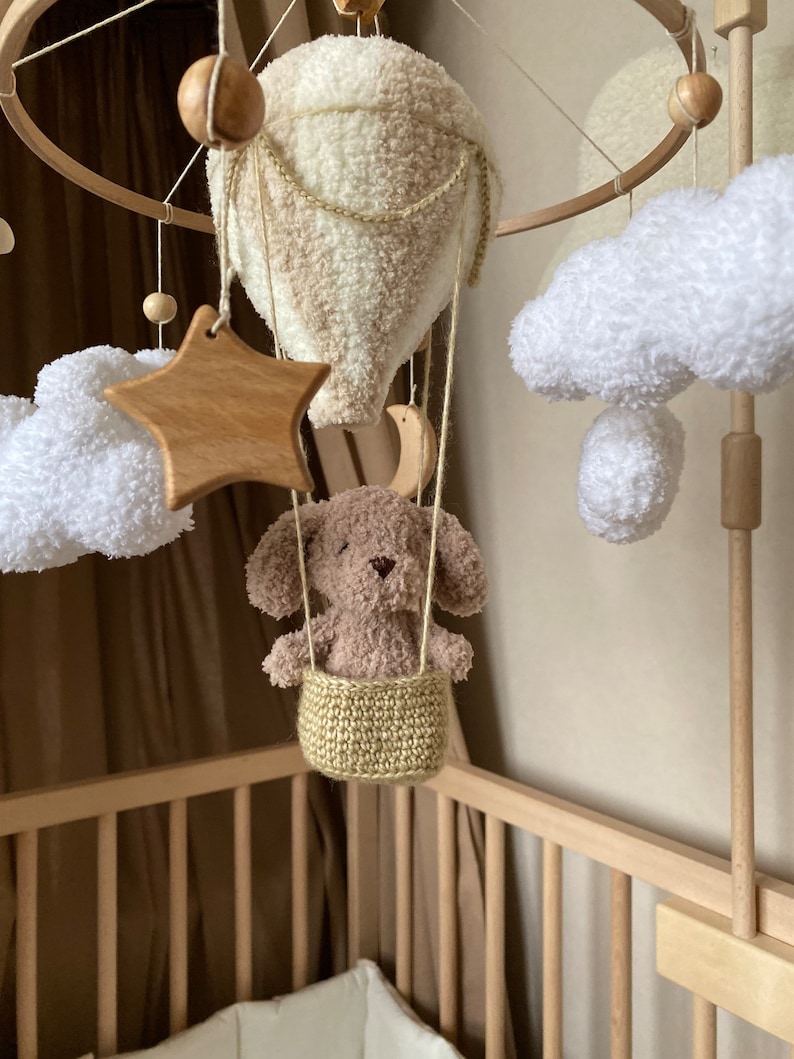 May include: A wooden baby mobile with a crocheted hot air balloon, clouds, and a star. A brown plush puppy sits in a crocheted basket hanging from the balloon.