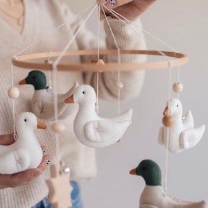 May include: A wooden baby mobile with a circular frame and five hanging elements. The elements include four plush duck figures, two with green heads and two with white heads, and a wooden star. The ducks are white with brown beaks and eyes. The mobile is suspended from a white string.