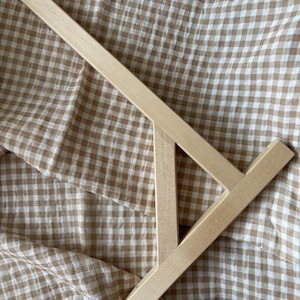 Puede incluir: Una herramienta de madera para hacer crepes, con un mango largo y un esparcidor en forma de T. La herramienta está hecha de madera de color claro y está colocada en diagonal sobre un fondo de tela a cuadros beige y blanco.