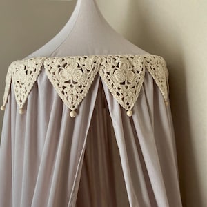 Pu&ograve; includere: Un baldacchino lilla chiaro con un bordo floreale all'uncinetto color crema. Il baldacchino ha una forma conica ed &egrave; realizzato in un tessuto morbido e fluido. Piccole perline di legno pendono dal bordo. Ideale per una cameretta o una camera da letto.