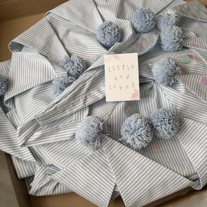 May include: A light blue and white striped fabric item featuring multiple light blue yarn pom-poms and scattered pastel confetti. A white rectangular card is placed on top with the text "little and loved" printed in a simple, lowercase font.