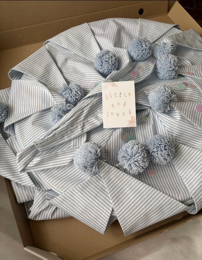 May include: A light blue and white striped fabric item featuring multiple light blue yarn pom-poms and scattered pastel confetti. A white rectangular card is placed on top with the text "little and loved" printed in a simple, lowercase font.